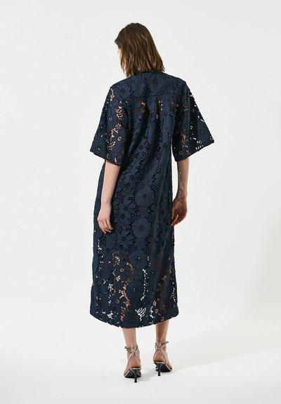Femme portant une robe midi en dentelle fleurie bleu marine avec des manches larges et des sandales noires à talons hauts avec des lanières, debout de dos.
