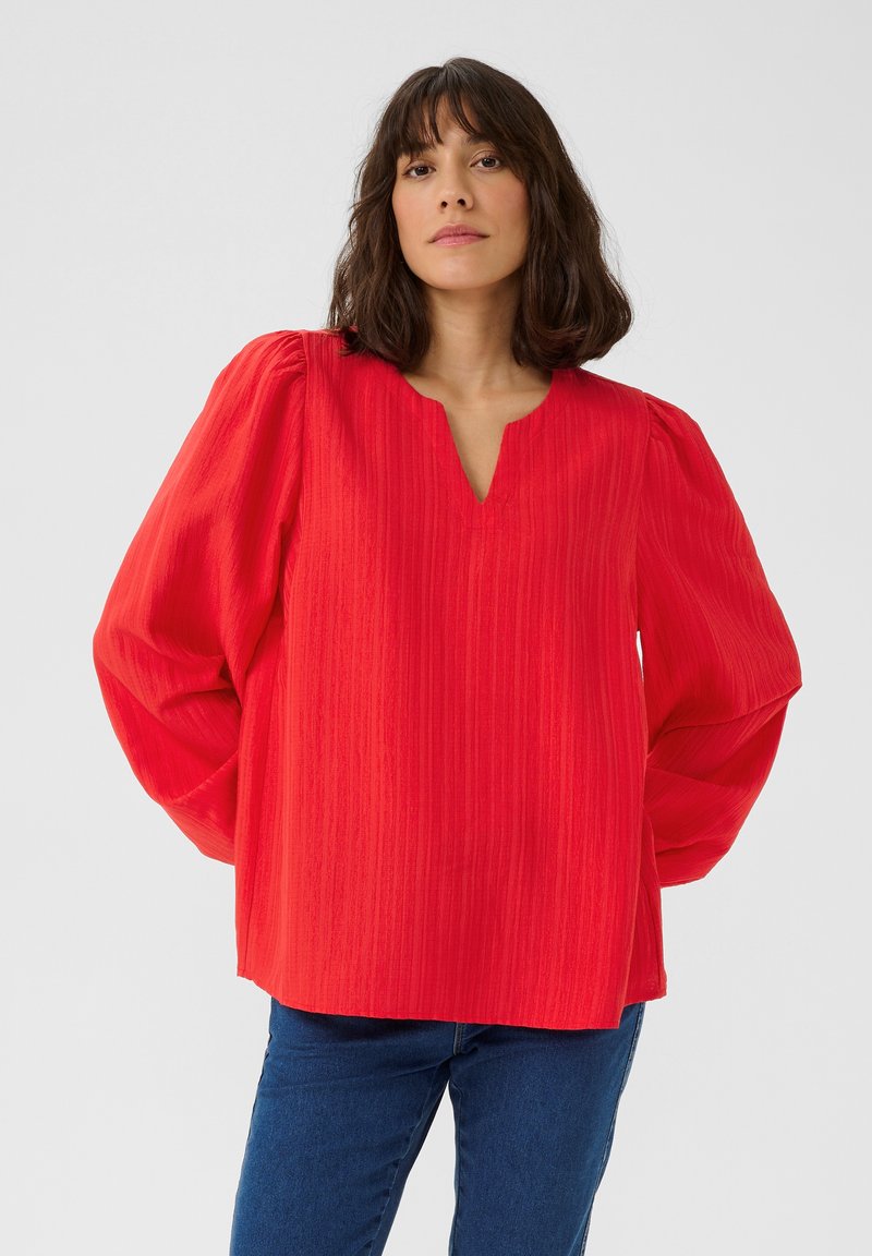 Blouse rouge texturée avec un décolleté en V, des manches bouffantes et une coupe décontractée, associée à un jean bleu pour un look décontracté.