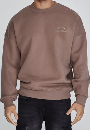 Hombre con sudadera marrón oversize con texto blanco pequeño en el pecho izquierdo y pantalones negros sobre un fondo liso.