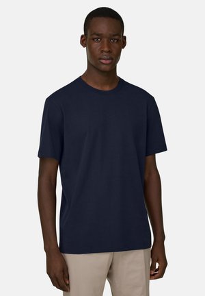 Joven vestido con una camiseta de manga corta azul marino lisa y pantalones beige, de pie frente a un fondo gris claro.