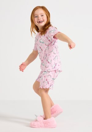 Hymyilevä punatukkainen tyttö, joka pitää yllään vaaleanpunaiset yksisarviskuosiset pyjamat ja pörröiset vaaleanpunaiset yksisarvissisäkengät, seisoo ja tasapainottelee ojennetuin käsin.