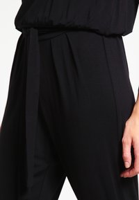 Černý, lehký overal s volným střihem, opaskem v pase a měkkou texturou. Obsahuje sklady podél boků.