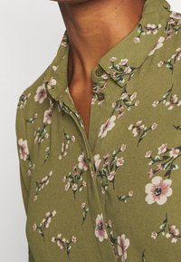 Groen bloemenoverhemd met een kraag, knoopdetails en een zachte textuur, versierd met roze en blauwe bloemenpatronen.