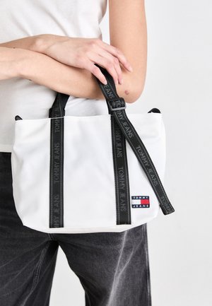 Osoba trzyma białą torbę typu tote Tommy Jeans z czarnymi paskami z logo, ubrana w biały top i ciemnoszare spodnie.