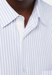 Záběr na límeček a horní část košile na knoflíčky s světle modrými a bílými vertikálními pruhy a detailem kapsy.