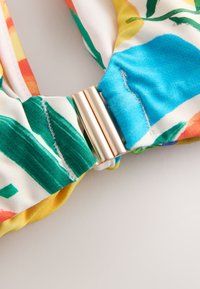 Tissu coloré avec des motifs verts, bleus, jaunes et orange, rassemblé et fixé par un petit fermoir cylindrique doré et brillant.