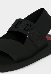 Μαύρο σανδάλι χωρίς λουράκια με δύο φαρδιές ελαστικές λουρίδες, η μία με ανάγλυφο το γράμμα «MOSCHINO», και επίπεδη σόλα με κόκκινη λεπτομέρεια καρδιάς στο εσωτερικό.