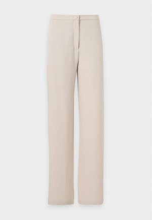 Femme portant un chemisier noir à manches longues avec volants et un col en V profond, et un pantalon large beige, debout sur un fond blanc.