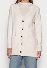 Beige pletený kardigan s výstřihem do V, šesti zlatými knoflíky, dvěma předními kapsami a žebrovanými manžetami a lemem. Měkká textura s volným střihem.