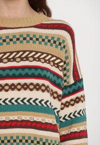 Gros plan d'un pull en tricot multicolore avec des motifs géométriques en beige, marron, bleu sarcelle, rouge et crème, porté par une personne aux cheveux châtain.