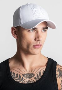 Bijela bejzbol kapa s utisnutim logotipom sprijeda, zakrivljenim vizirima i podesivim remenom. Model nosi crni top bez rukava s vidljivim tetovažama.