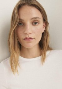 Nahaufnahme eines Porträts einer Person mit langen, welligen braunen Haaren und heller Haut, die ein weißes geripptes Shirt vor einem neutralen Hintergrund trägt.