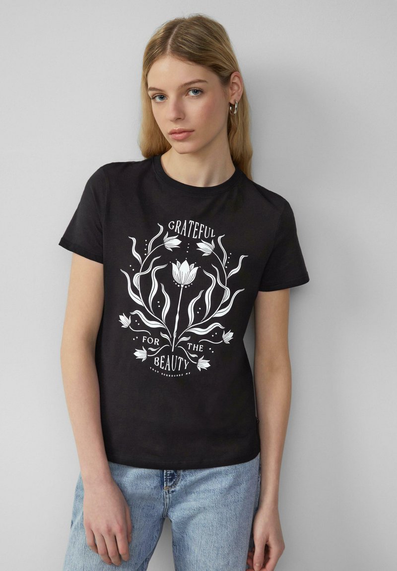 Zwarte katoenen T-shirt met korte mouwen, voorzien van een witte bloemmotief en de tekst "DANKBAAR VOOR DE SCHONE DINGEN" in een decoratief lettertype.