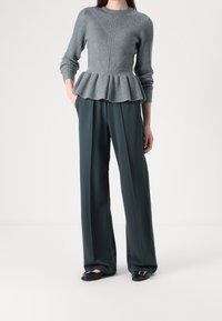 Femme portant un pull péplum gris texturé à manches longues, un pantalon large foncé, et des ballerines noires pointues avec boucles.