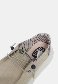 Chaussure en tissu beige avec une semelle claire, dotée d'un intérieur en rose doux à motifs et d'accents bleus. Comprend des lacets élastiques et une petite étiquette de logo.