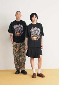 Black graphic T-shirts with bold front designs, paired with camouflage pants and a pleated black skirt. Brown shoes and white socks visible.