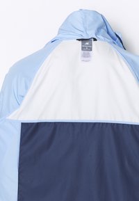 Kapotová bunda s ľahko modrými, bielymi a námorníckymi farebnými blokmi, zobrazená zo zadnej strany s viditeľnou nálepkou New Balance vo vnútri goliera.