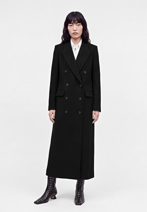 Femme debout portant un long manteau noir croisé, une chemise blanche et des bottines noires à lacets sur un fond uni.