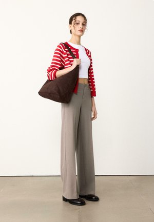 Rjava torbica iz semiša v obliki hobo torbe, ki jo drži oseba v rdeče-beli črtasti jopi, belem kratkem topu in sivih širokih hlačah s črnimi čevlji.