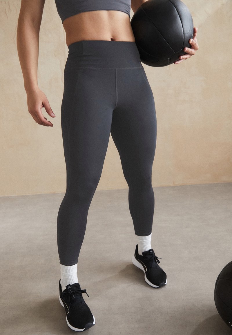 Szare legginsy sportowe z wysokim stanem, wykonane z elastycznego materiału, w połączeniu z czarnymi butami sportowymi i białymi skarpetkami do kostek.