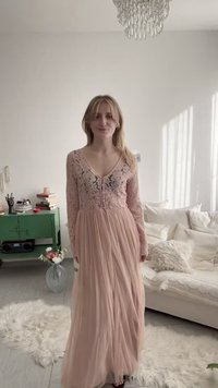 Giovane donna in un lungo vestito di pizzo rosa, in piedi in un luminoso soggiorno con un divano bianco, un armadio verde e un lampadario sopra.