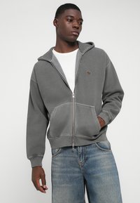 Grauer Reißverschluss-Hoodie mit Kapuze und Kordelzug, Kängurutaschen und einem kleinen Logo. Über einem weißen T-Shirt getragen und mit hellblauen Jeans kombiniert.