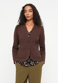 Frau mit lockigem Haar, die ein braunes Blazer über einer gemusterten Bluse und olivgrünen Hosen trägt, steht vor einem schlichten weißen Hintergrund.