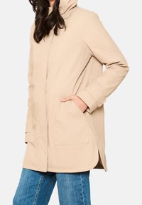 Beżowy wodoodporny płaszcz z kapturem, zapięciem na guziki i kieszeniami z przodu. Posiada luźny krój i gładką fakturę.