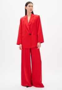 Conjunto de pantalones rojos que consiste en un blazer largo con cierre de botón y pantalones de pierna ancha, confeccionado en una tela suave y estructurada.