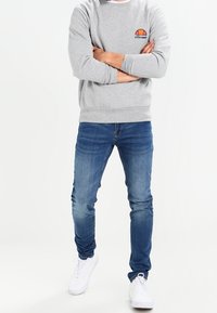 Sudadera gris con cuello redondo y un pequeño logotipo de color en el pecho, combinada con jeans azul ajustados y zapatillas blancas.