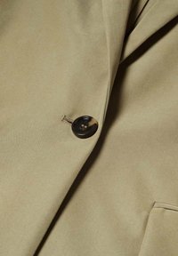 Gros plan du revers beige d'un manteau avec un seul bouton foncé marbré et une partie d'une poche avant visible.
