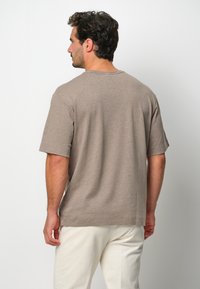 Brązowa koszulka z krótkim rękawem, wykonana z miękkiego materiału, o luźnym kroju, okrągłym dekolcie i subtelnej teksturze. Noszona z jasnymi spodniami.