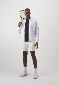 Chaqueta blanca de Nike sobre una camiseta negra, combinada con pantalones cortos blancos. Sosteniendo una raqueta de tenis y una pelota de tenis verde brillante. Zapatillas blancas de Nike.
