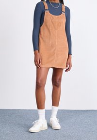 Latzkleid aus Cord in Hellbraun, mit verstellbaren Trägern, einer Fronttasche, kombiniert mit einem navyblauen Langarmshirt und weißen Sneakers.