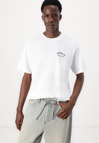 Hvid, kortærmet t-shirt med et sort logo-detalje på venstre bryst. Sammen med lyseblå, løstsiddende denimjeans.