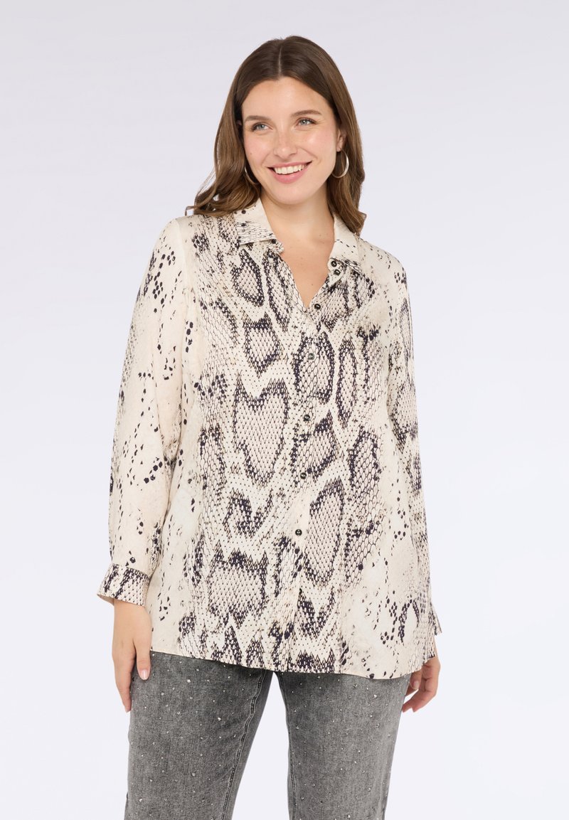 Donna sorridente, che indossa una camicia a maniche lunghe beige e nera con stampa serpente e jeans grigi con decorazioni in perle.
