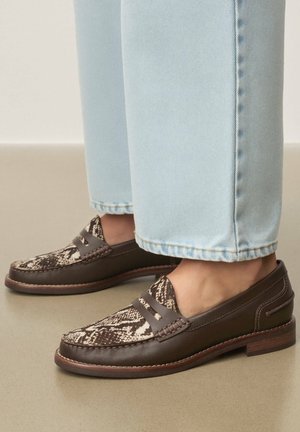 Barna bőr mokaszin kígyómintás szövettel, világoskék farmerrel viselve, bézs padlón.