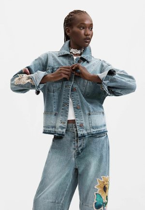 Frau mit Zöpfen knöpft eine Jeansjacke zu, trägt dazu passende Jeans mit einem Sonnenblumen- und Schmetterlingsaufnäher, vor einem schlichten Hintergrund.