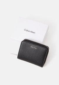 Black textured leather wallet with a silver zipper, featuring a logo on the front, placed beside a white box with a brand name.