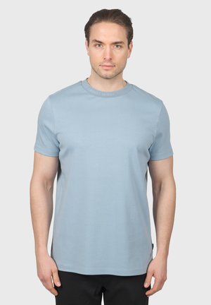 Hombre con cabello castaño vistiendo una camiseta de manga corta lisa azul claro y pantalones negros, de pie frente a un fondo gris claro.