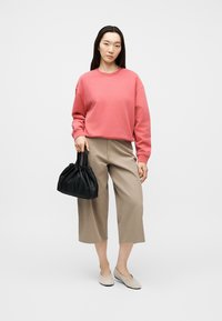Femme portant un sweat-shirt corail et un pantalon beige large court, tenant un sac à main noir, debout devant un fond clair uni.