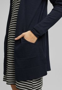 Marine gebreide cardigan met lange mouwen, voorzakken en geribde zoom, over een gestreepte jurk in zwart en wit.