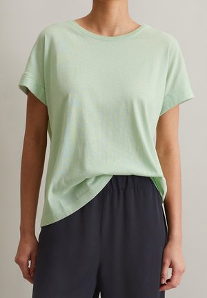 Jasnozielona koszulka z krótkim rękawem wykonana z miękkiego materiału, o luźnym kroju i zaokrąglonym dołu, zestawiona z ciemnymi spodniami.