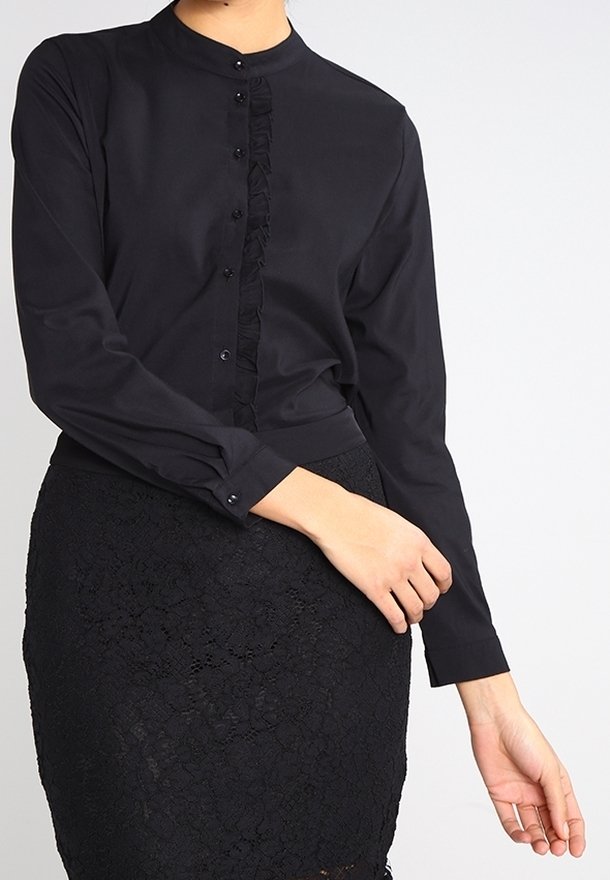 Blouse noire à manches longues avec col mandarin, patte de boutonnage à volants et boutons assortis. Associée à une jupe noire en dentelle.