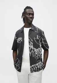 Homem com dreadlocks curtos, vestindo uma camisa preta de mangas curtas com linhas abstratas brancas sobre uma t-shirt branca e calças de cor clara.