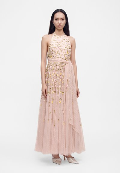 Femme portant une robe halter rose sans manches avec broderies florales, une ceinture nouée à la taille et des talons pointus assortis, debout devant un fond blanc.