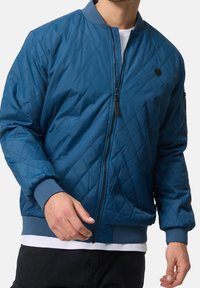 Man draagt een blauwe doorgestikte bomberjack met ritssluiting aan de voorkant, geribbelde boorden en zoom, en een klein rond zwart logo op de borst.