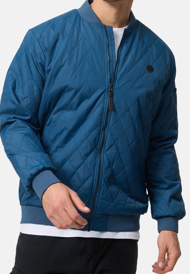 Man draagt een blauwe doorgestikte bomberjack met ritssluiting aan de voorkant, geribbelde boorden en zoom, en een klein rond zwart logo op de borst.
