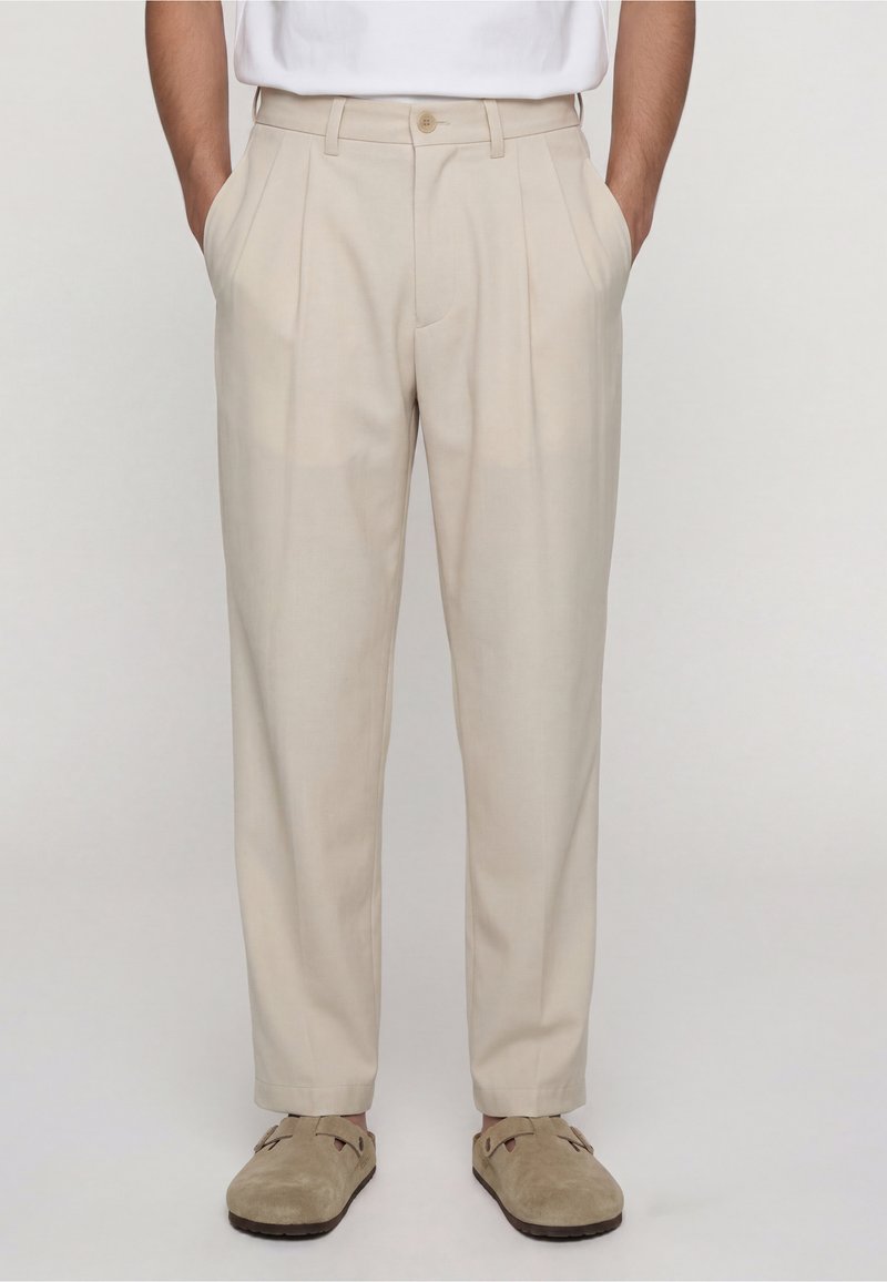 Homme portant un pantalon plissé beige, les mains dans les poches, assorti à des chaussures en daim beige à enfiler et une chemise blanche rentrée.