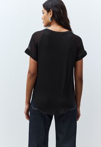 Femme aux cheveux foncés portant des boucles d'oreilles argentées, un top noir à manches courtes avec des manches transparentes et un jean en denim foncé, debout de dos.